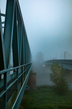 View of a metal bridge structure extending into thick morning fog with a secondary bridge visible in the distance over the riverbank in Mannheim, Baden-Wurttemberg, Germany.