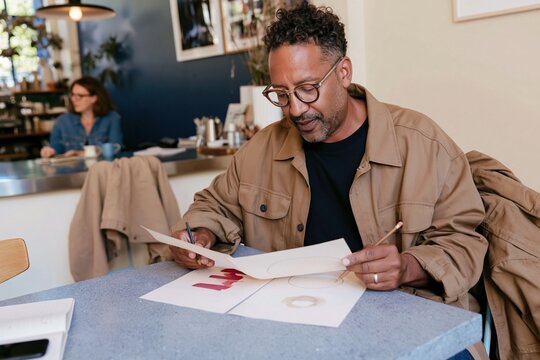 Black man comparing two blank printed sheets on a cool grey table, a natural scene in a lived-in office for copywriting and freelance decision making