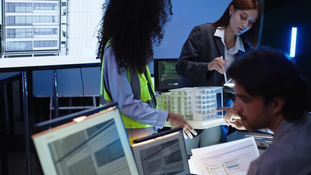 Female manager holding digital tablet while pointing at architectural building model during construction planning session with engineering team reviewing project documents at desk