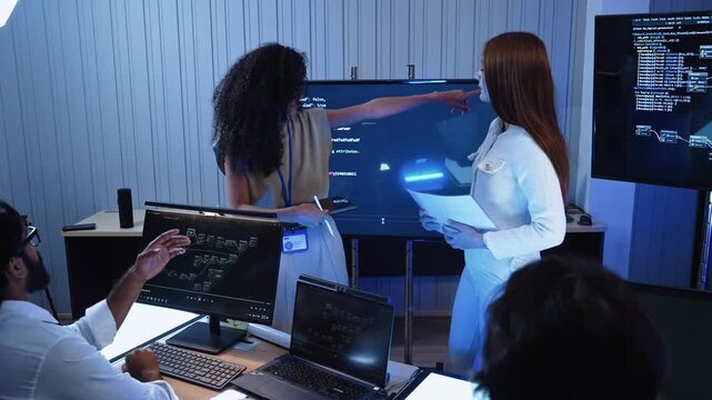 Female tech leader pointing at big display screen while presenting software code structure to programming team working on computer workstations during office development meeting