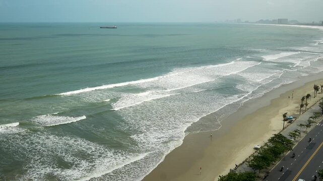 Elevated view of Da Nang coastline shows ocean waves reaching a sandy beach beside a palm-lined promenade and coastal road, with light traffic and a cargo ship offshore under hazy daylight.