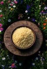Obraz premium Healthy nutrition and organic food, bowl of raw couscous grains on a rustic wooden stump surrounded by a colorful summer meadow with wildflowers