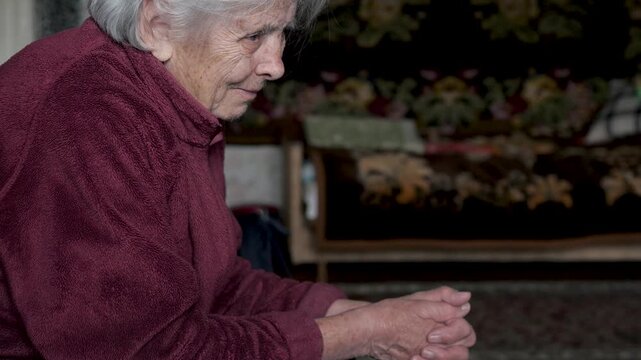 Lonely senior woman with grey hair sitting in dark room. Old granny wrinkled hands clasped as reflects on memories and experiences, portraying themes of nostalgia, aging and solitude