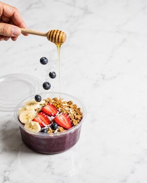 Hand drizzling honey on healthy acai smoothie bowl with falling blueberries on white marble