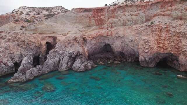 Drone shot of dramatic rocky cliffs and natural sea caves along the clear turquoise shore at Tsigrado Beach on Milos Island, Greece