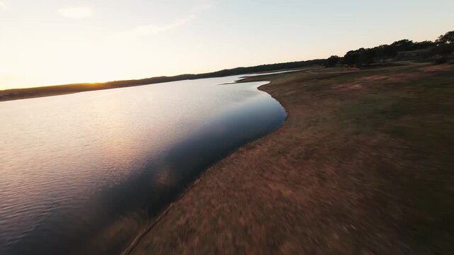 Golden sun dips low over calm lake surface. Water ripples gently as sky softens to twilight hues. Dry shoreline curves with sparse trees in fading light. Stillness blankets the scene, peaceful