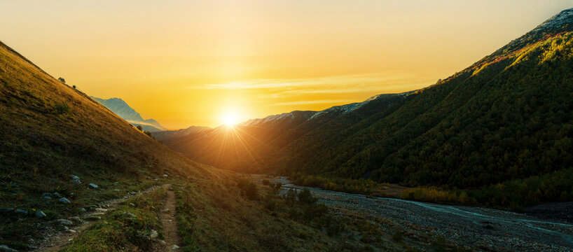 Panoramic landscape photograph of glowing golden sunrise with sunstar over mountain valley and trekking path near Adishi Glacier in Svaneti Georgia.