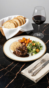 Grilled filet mignon with special truffle sauce, brown rice, baby carrots, fresh salad, a bread basket, and a glass of red wine on an elegant marble table
