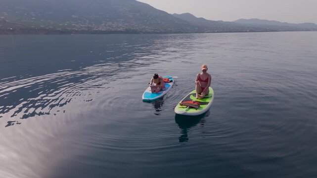 Two Women Stretch on SUP Boards as One Performs Seated Forward Bend and Friend Waves at Drone on Calm Sea Near Kalamata, Greece