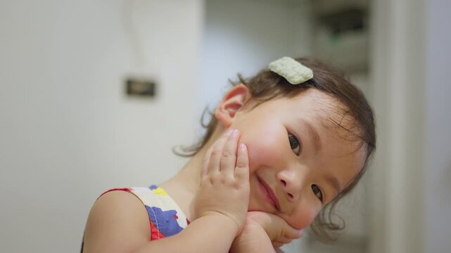 A cute Asian toddler girl holds her face with both hands and smiles gently, looking away with a sweet expression of childhood wonder and contentment in her home.