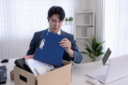 Young Asian businessman packing documents and personal items in cardboard box after resignation in modern office