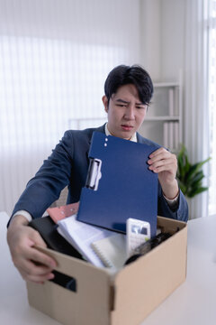 Young Asian man holding folder examining belongings in cardboard box after job termination in bright office with copy space