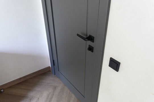 matte‑gray interior door features sleek black lever handle and matching lock. minimalist black wall switch, light wood herringbone floor, clean white walls complete look