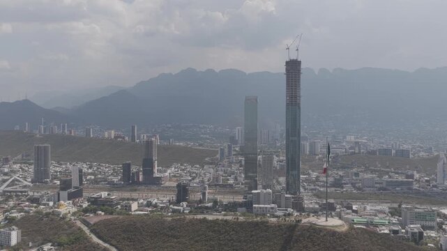 Aerial panoramic view, drone flying sideways with views of the Mexican Flag at Obispado with Torre Rise and the San Pedro Skyline in the background. Shot in D-Log M