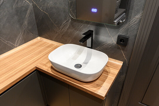 modern bathroom features white vessel sink on  warm wooden countertop. dark marble walls, black faucet, mirrored cabinet with subtle lighting create sleek look