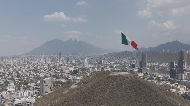 Aerial view of the monumental Mexican flag waving atop Cerro del Obispado with Cerro de la Silla and the city skyline in Monterrey, Nuevo Leon, Mexico, D-Log M