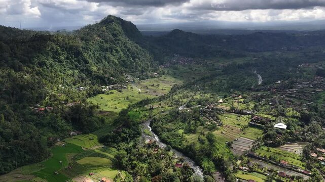 Aerial view of lush Mount Agung foothills with winding road and river under dramatic overcast sky showing shifting cloud shadows and sunlit valley patches, with mountain, foothill, valley, road detail