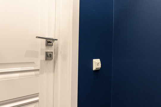 White paneled door with brushed‑metal lever handle and lock contrasts deep blue wall. minimalist white wall switch adds subtle accent