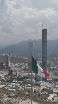 Aerial view of the monumental Mexican flag waving with Torre Rise, the tallest building in Latin America, behind it in Monterrey, Nuevo Le&oacute;n. Vertical video. D-Log M
