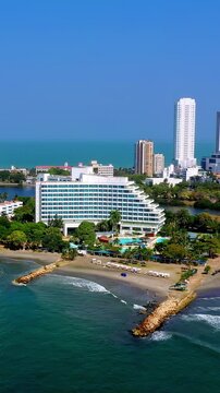 Aerial view of a luxury hotel resort on the beach in Cartagena, Colombia, with tropical trees, swimming pools, and modern skyscrapers in the background