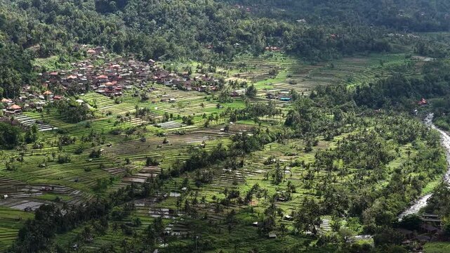 Aerial panorama of verdant Mount Agung foothills with village, terraced fields, and winding river under shifting sunlight, with foothill, valley, river, stream detail