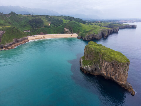 Aerial coastal view from the Atlantic Ocean overlooking calm waters, a rugged rocky islet, Playa de Andr&iacute;n beach, lush green headlands, and cloud-covered mountains. Costa Verde, Asturias, Spain.