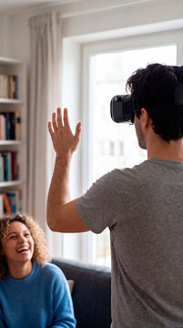 Man wearing VR headset waves at laughing woman seated on cozy living room sofa