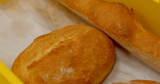close-up view of baked round loaves and baguette resting in bright yellow perforated plastic crate inside bakery or kitchen setting, warm overhead lighting, with