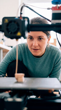 Woman crouches watching 3D printer carefully build small ceramic shape