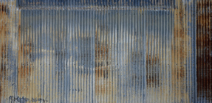 Blue tinted corrugated galvanized metal surface with rust showing a blue upper band, distressed rusted side panels, thin tan paint band, and repair patch in a horizontal close-up composition