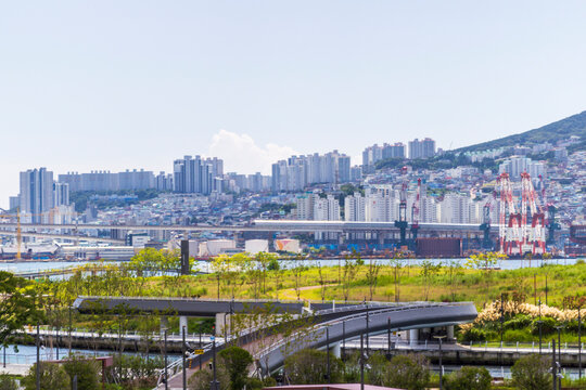 A dense city skyline with apartment towers, construction cranes, elevated infrastructure, waterfront greenery, and a wooded hillside beneath an expansive sky.