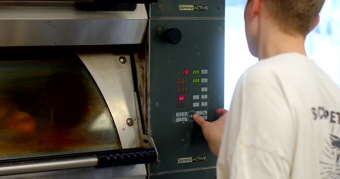young bakery worker presses buttons on digital control panel of industrial oven while loaves bake behind smudged viewing glass, scene shows stainless steel oven