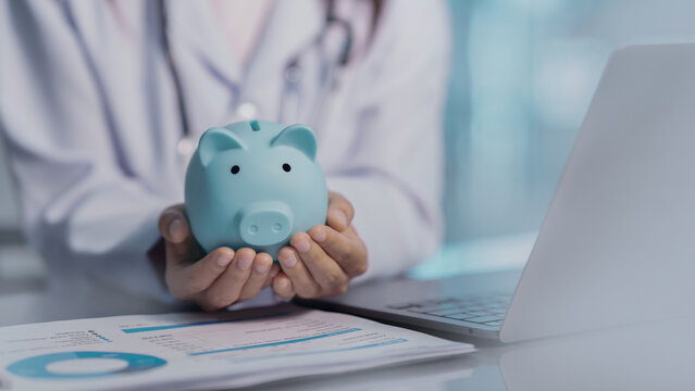 Doctor holding blue piggy bank sitting at desk with laptop and medical report. Professional healthcare worker focused on financial planning, personal savings, insurance policy, and money management.