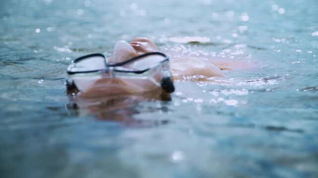 A child wearing swimming goggles relaxes while floating on his back in a natural lake. The footage captures summer recreation, open-water swimming, and carefree childhood.