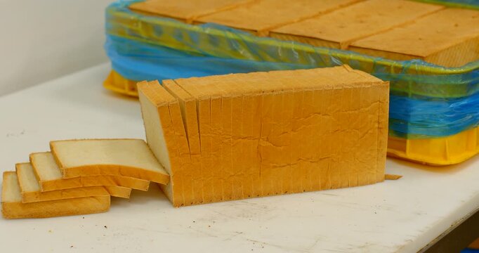 multiple pre-sliced white sandwich loaves placed on worktable showing neat vertical cut marks and separated slices arranged in foreground, stacked yellow trays bread