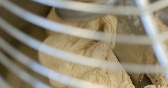 dough being gathered and folded around large metal paddle inside commercial stainless steel mixing bowl, visible through curved protective guard wires, warm, with