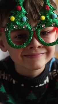 Vertical video. Christmas holiday. Closeup portrait of joyful child wearing funny Christmas eyeglasses at home