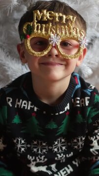 Vertical video. Christmas holiday. Joyful cute child wearing funny Christmas eyeglasses at home