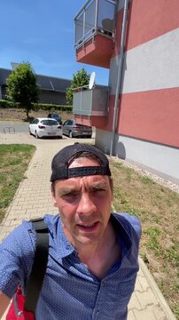 close-up selfie of adult man walking toward camera in sunny residential parking area, wearing backward baseball cap, blue patterned short-sleeve shirt, and, with
