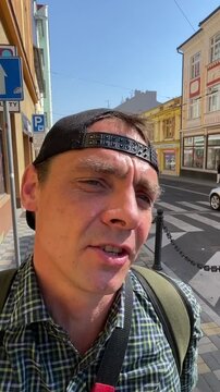 close-up selfie of man in checked shirt and backward mesh cap talking to camera while standing on sunlit urban sidewalk, directional signage visible in background