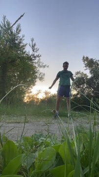 adult male standing on uneven dirt track inside tree-lined grassy clearing at dusk, raising both arms overhead in open, celebratory pose, scene captured from, with