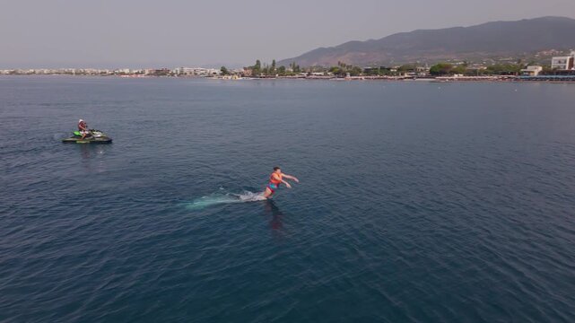 Beginner Woman Practices Flyboard Takeoff and Balance Near Water Surface While Jet Ski Operator Supervises in Kalamata, Greece &ndash; Aerial View