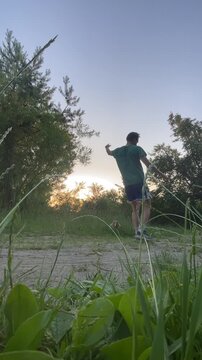 adult male in green t-shirt and blue athletic shorts performs brief leg raise, bends forward, then straightens up on sandy path by grassy field during sunset, low