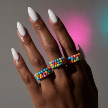 Woman's hand with white nails wearing colorful beaded rings against a gray background with pink and blue light