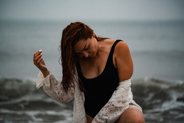 Woman in black swimsuit stands by the shore at the beach during cloudy weather. A woman wades in shallow water at the beach on a cloudy day. She looks down at the waves. © Alisa