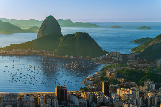 Panoramic view from the Dona Marta viewpoint looking out over the Rio de Janeiro cityscape, Botafogo Bay, and Sugarloaf Mountain at sunset in Brazil.