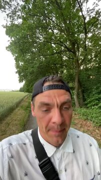close-up handheld selfie of adult male speaking while walking on narrow rural path beside crop field and dense tree line, head tilted and mouth open indicating man