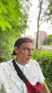 selfie-style outdoor motion scene of man walking narrow leafy path, wearing backward cap, patterned shirt, and red crossbody bag