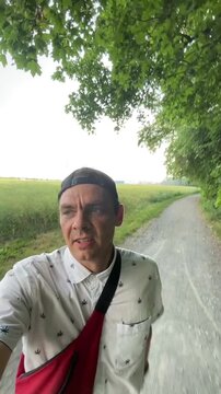 man walking along shaded forest trail with backward mesh cap, white short-sleeve patterned shirt, and red crossbody bag, mouth slightly open and eyes directed man