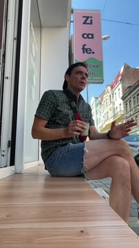 male-presenting adult seated on wooden bench outside cafe, lifting disposable cup toward mouth while gesturing with free hand and gazing upward, low-angle vertical